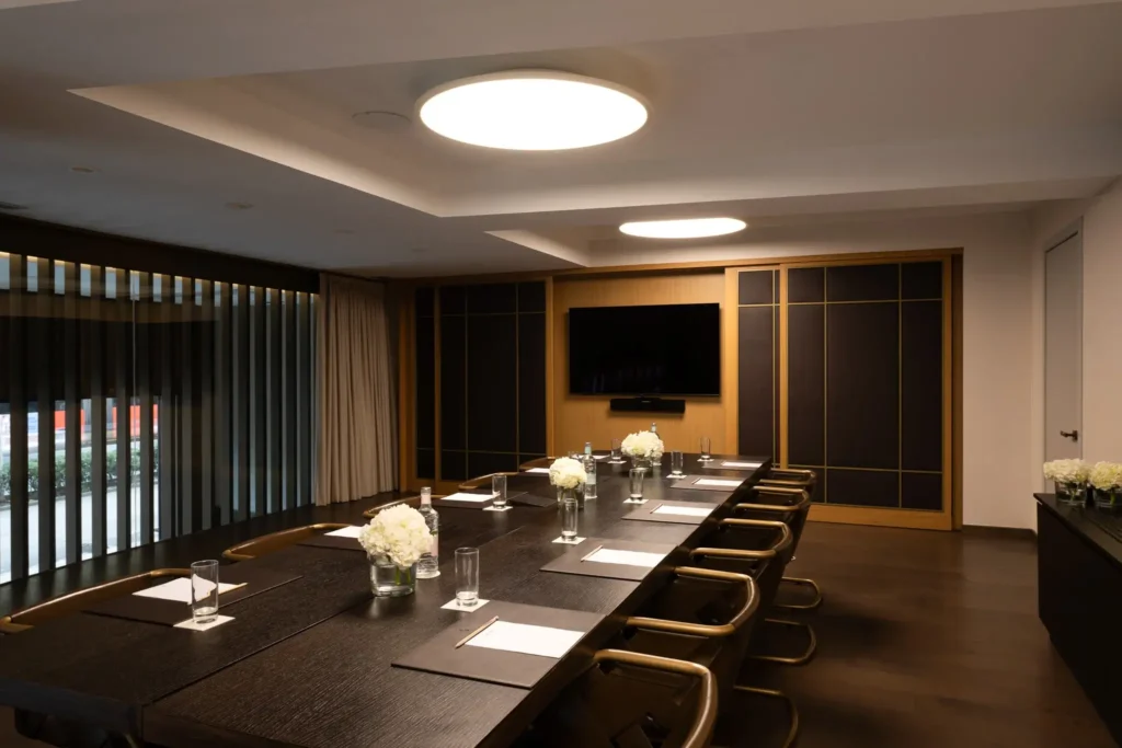 A modern conference room with a long table, chairs, notepads, glasses of water, flower centerpieces, a wall-mounted TV, and ceiling lights.